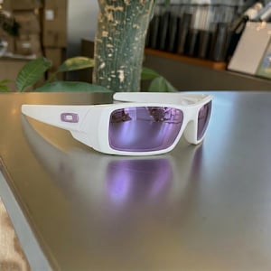 May include: White sunglasses with purple mirrored lenses. The sunglasses have a rectangular frame and a small, purple logo on the side. The lenses reflect the surrounding environment.