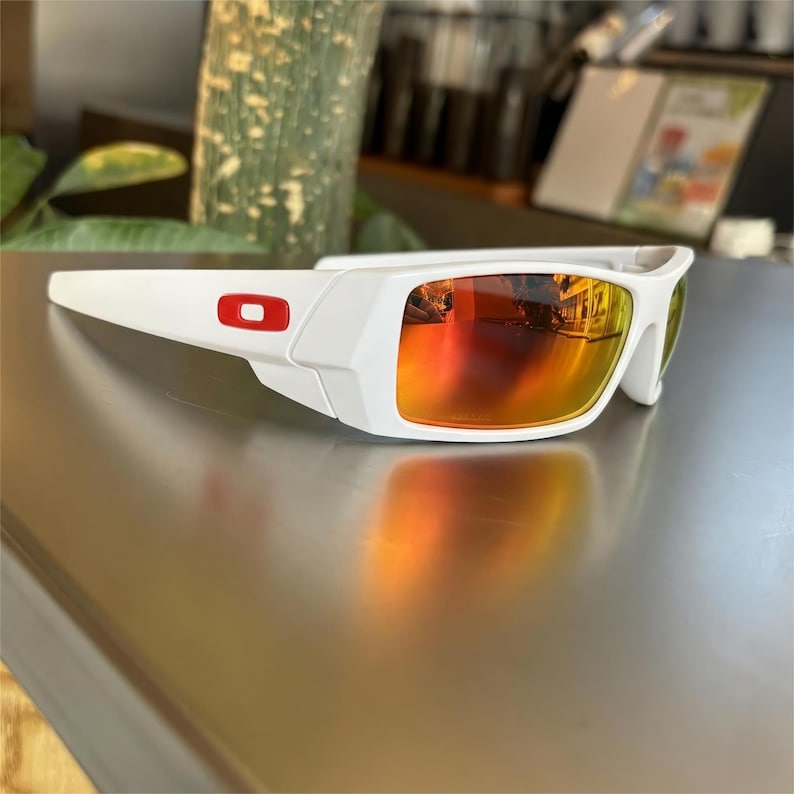 May include: White sunglasses with a red logo on the frame and orange-yellow mirrored lenses. The sunglasses are resting on a reflective surface. The lenses reflect the surrounding environment.