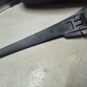 May include: A close-up of a black, angular object with the word "GASCAN" printed on it. The object has a long, slender arm and a rectangular section at the end. The object is on a light-colored surface.