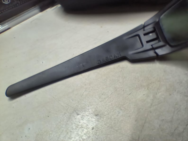 May include: A close-up of a black, angular object with the word "GASCAN" printed on it. The object has a long, slender arm and a rectangular section at the end. The object is on a light-colored surface.