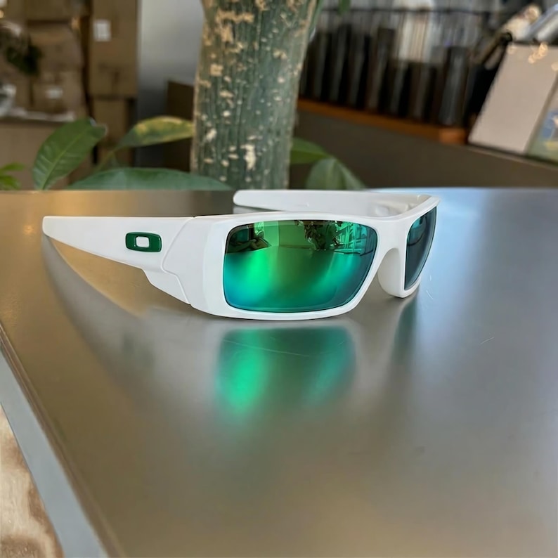 May include: White sunglasses with a green mirrored lens. The sunglasses have a rectangular frame with a green logo on the side. The lenses reflect the surrounding environment.