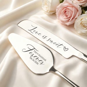 May include: Silver cake knife and server set with the words "Love is sweet" and "Forever" engraved. The server is personalized with a name and date. The set is displayed on a white satin surface with roses.