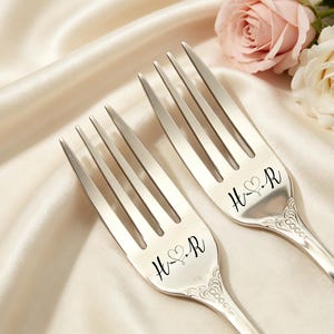 May include: Two silver forks engraved with the initials "H & R" and a heart symbol, set on cream satin. Pink and white roses are in the background, suitable for a wedding or anniversary celebration.