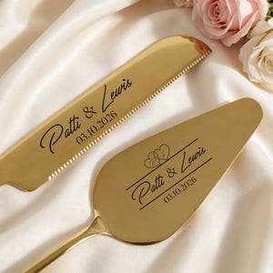 May include: Gold-colored wedding cake knife and server set. The knife has a serrated edge and the server is spade-shaped. Both are engraved with "Patti & Lewis" and the date 03.10.2026. The set is displayed on a white satin surface.
