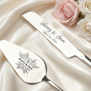 May include: A silver cake knife and server set. The knife is engraved with "Avery & Sam" and "April 26, 2025". The server is engraved with "Avery & Sam" and "November 26, 2025" along with floral designs. The set is displayed on a white satin surface.