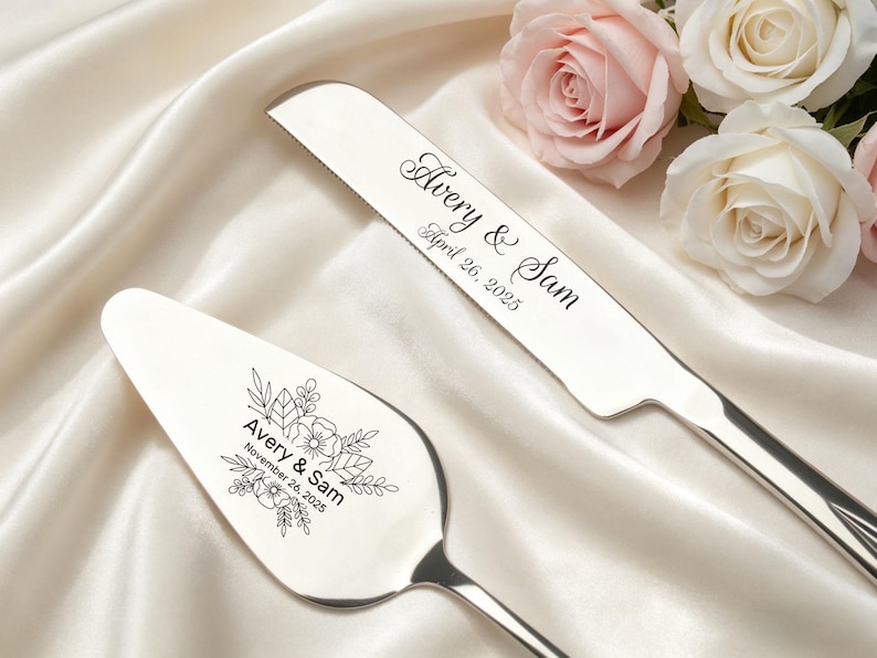 May include: A silver cake knife and server set. The knife is engraved with "Avery & Sam" and "April 26, 2025". The server is engraved with "Avery & Sam" and "November 26, 2025" along with floral designs. The set is displayed on a white satin surface.