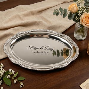 Peut inclure: Un plateau de service ovale de couleur argentée avec un bord décoratif en relief orné de perles et de motifs floraux. Le centre du plateau présente le texte gravé Teague & Lacey et la date October 15, 2026, en police script et serif.