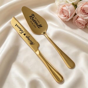 May include: A gold-colored cake knife and server set with engraved text. The knife reads "Always & Forever" and the server reads "Mr. & Mrs. Russell 03.06.2026". The set is displayed on a white satin surface, with roses in the background.