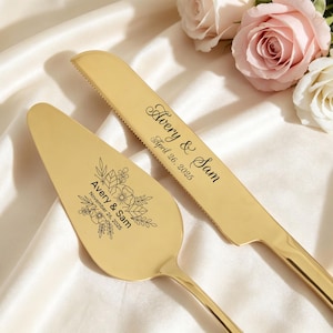May include: Gold-colored wedding cake knife and server set. The knife and server are engraved with the names "Avery & Sam" and the wedding date "April 26, 2025" on the knife and "November 26, 2025" on the server. The set is displayed on a white satin surface with roses.