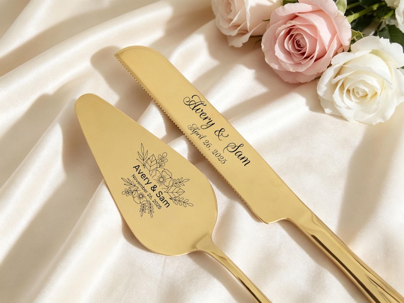 May include: Gold-colored wedding cake knife and server set. The knife and server are engraved with the names "Avery & Sam" and the wedding date "April 26, 2025" on the knife and "November 26, 2025" on the server. The set is displayed on a white satin surface with roses.