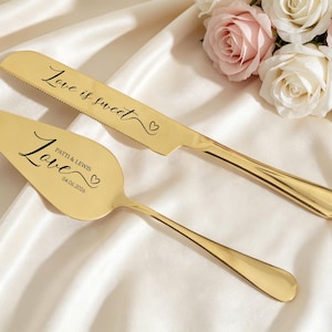 May include: Gold-colored cake knife and server set with the words "Love is sweet" and "Love" inscribed. The server is personalized with names and a date. The set is displayed on a white satin surface, with a bouquet of roses in the background.