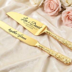 Könnte beinhalten: Ein goldfarbenes Tortenmesser- und Tortenheber-Set mit verzierten, floral geprägten Griffen. Der Tortenheber ist mit den Namen Lea & Daniel und dem Datum 09.24.2026 graviert, während das gezackte Messer die Inschrift together forever trägt.