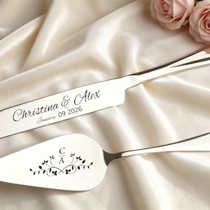 May include: Silver cake knife and server set. The knife is engraved with "Christina & Alex" and the date "January 09 2026". The server features the initials "C A" with floral accents. The set is displayed on a cream-colored satin cloth.