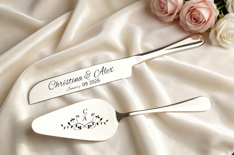 May include: Silver cake knife and server set. The knife is engraved with "Christina & Alex" and the date "January 09 2026". The server features the initials "C A" with floral accents. The set is displayed on a cream-colored satin cloth.