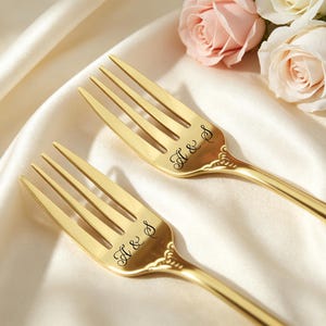 May include: Two gold-toned metal forks featuring a polished finish and ornate detailing at the neck. Each fork is engraved with the letters A & S in a cursive script font. The forks are positioned diagonally, showcasing their metallic lustre.