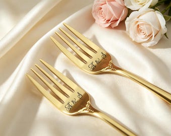May include: Two gold-toned metal forks featuring a polished finish and ornate detailing at the neck. Each fork is engraved with the letters A & S in a cursive script font. The forks are positioned diagonally, showcasing their metallic luster.