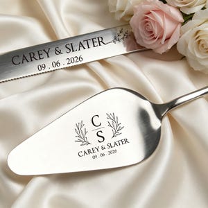 May include: A silver cake knife and server set with engraved text. The knife reads "CAREY & SLATER" and "09.06.2026". The server features the initials "C" and "S" with "CAREY & SLATER" and "09.06.2026". The set rests on a white satin surface.