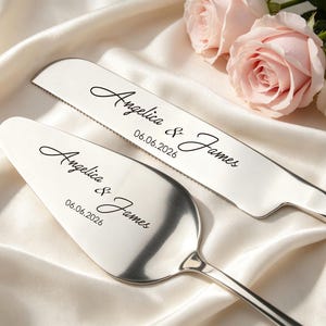 Puede incluir: Un juego de cuchillo y servidor de pastel plateado con los nombres "Angelica & James" y la fecha "06.06.2026" grabados en escritura negra. El juego descansa sobre una superficie de satén blanco, con rosas rosas y blancas.