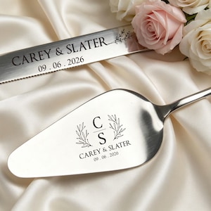 May include: A silver cake knife and server set with engraved text. The knife reads "CAREY & SLATER" and "09.06.2026". The server features the initials "C" and "S" with "CAREY & SLATER" and "09.06.2026". The set rests on a white satin surface.