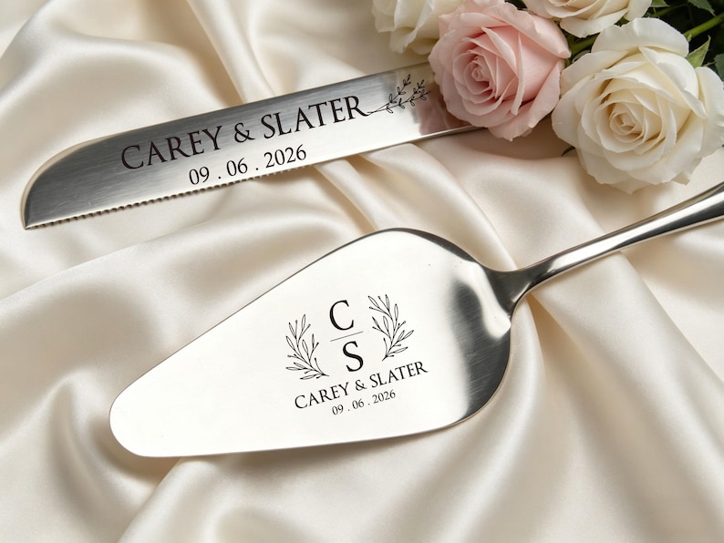 May include: A silver cake knife and server set with engraved text. The knife reads "CAREY & SLATER" and "09.06.2026". The server features the initials "C" and "S" with "CAREY & SLATER" and "09.06.2026". The set rests on a white satin surface.