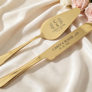 May include: A gold-colored cake server and knife set. The server has a wide, flat blade, and the knife has a serrated edge. Both are engraved with the names "Carey & Slater" and the date "09.06.2026". The set rests on a white fabric with pink and white roses.