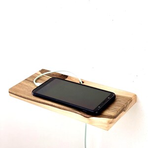 May include: A rectangular wooden wall shelf with a natural grain pattern and a dark edge, featuring a circular cutout for a charging cable. A black smartphone rests on the surface, connected to a white charging cable that passes through the hole.