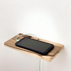 Könnte beinhalten: Ein kleines Holzregal mit natürlicher Holzmaserung, an einer weißen Wand befestigt. Ein schwarzes Smartphone liegt auf dem Regal, mit einem hellgrünen Ladekabel. Das Regal hat ein Loch für das Kabel.