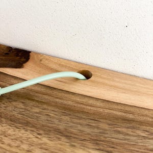 Puede incluir: Una superficie de madera clara con un patrón de veta natural y un nudo oscuro en el lado izquierdo. Un orificio circular está perforado en la madera, a través del cual pasa un cable liso de color verde menta pálido, extendiéndose hacia afuera.