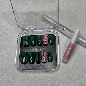 May include: A set of green and pink press-on nails in a clear plastic case. The nails are a glossy green, with two pink nails featuring black spots. A tube of nail glue is also included.