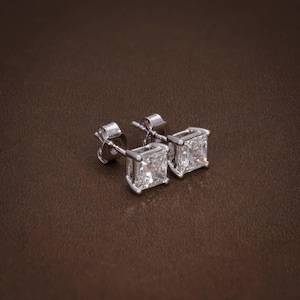 Puede incluir: Un par de pendientes de diamantes cuadrados con engaste de plata. Los pendientes se exhiben sobre una superficie marrón, mostrando su brillo y claridad. Los pendientes tienen un diseño clásico y elegante.