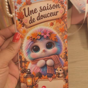 Peut inclure: Une couverture de carnet rectangulaire illustrée d'une créature duveteuse multicolore aux grands yeux, portant une écharpe rose et une couronne de fleurs. Le décor automnal inclut des feuilles, une lanterne et un écureuil. Texte : Une saison de douceur.