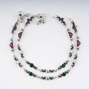 May include: A pair of silver anklets featuring a repeating pattern of round and teardrop-shaped red, green, and clear stones. Each anklet is constructed with linked metal segments and includes small dangling charms at the clasp closure ends.
