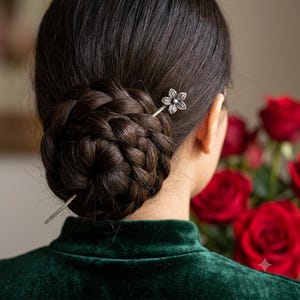 Könnte beinhalten: Eine silberfarbene Haarnadel aus Metall mit einem fünfblättrigen Blumendesign an einem Ende. Das Haaraccessoire steckt in einem dunkelbraunen geflochtenen Dutt, wobei das spitze Ende der Nadel auf der anderen Seite der Frisur sichtbar ist.