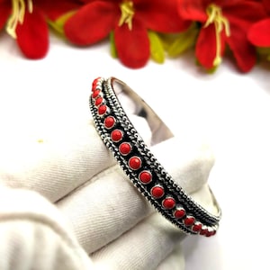 Puede incluir: Una pulsera de plata tipo brazalete con una fila central de pequeñas piedras redondas de color rojo brillante. La banda metálica está decorada con un borde texturizado de cuentas a ambos lados de las piedras, creando un acabado metálico detallado.