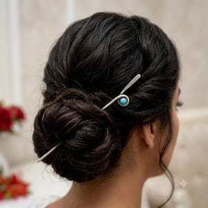 Op de afbeelding: Een zilverkleurige metalen haarspeld met een glad, langwerpig ontwerp dat in een cirkelvormige lus buigt. Het midden van de lus is geaccentueerd met een kleine, ronde, helder turquoise steen. De speld zit in een donkerbruine haarknot.