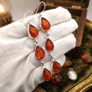 May include: A pair of long drop earrings featuring three teardrop-shaped amber stones set in silver. Each earring consists of three vertically linked amber pieces in a warm orange hue, suspended from a silver hook wire for pierced ears.
