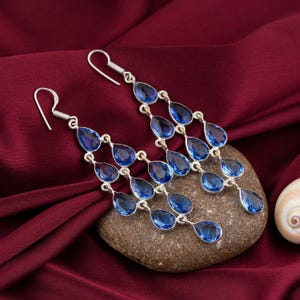 Puede incluir: Un par de pendientes largos tipo candelabro con múltiples piedras preciosas azules facetadas en forma de pera, engastadas en metal plateado. Cada pendiente consta de varias piedras colgantes en forma de lágrima en un diseño vertical.