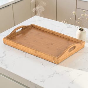May include: A rectangular bamboo serving tray with curved handles. The tray is light brown and has a smooth surface. It is sitting on a white countertop. A small vase with white flowers is in the background.