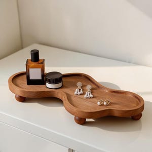 May include: A wooden tray with a unique, flowing shape, resting on four small, rounded feet. The tray holds a perfume bottle, a small jar, and a pair of earrings. The tray is made of a warm, medium-toned wood.