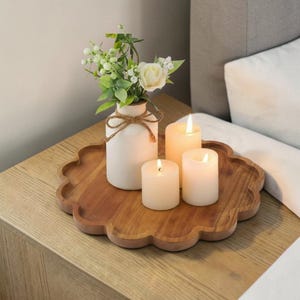 May include: A wooden, scalloped-edge tray holds a white vase with flowers and three lit candles. The vase is tied with twine. The candles are different sizes and have a warm glow. The tray sits on a wooden nightstand.