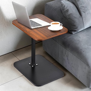 May include: A small, brown wooden side table with a black metal base and support. A laptop and a cup of coffee on a white saucer sit on the table. The table is positioned next to a gray sofa.