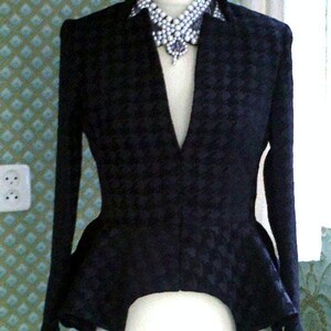 May include: A black blazer with a peplum hem and a pointed collar. The blazer has a floral pattern and is worn with a large pearl necklace.