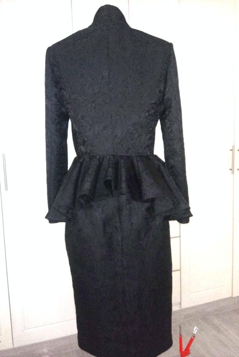 May include: A black dress with a high neck and a ruffled peplum. The dress is made of a textured fabric and has long sleeves.