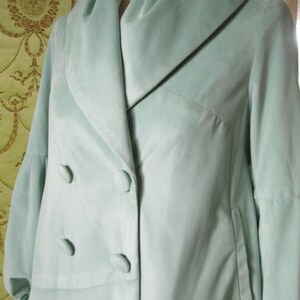 May include: A light blue velvet coat with a large collar and double breasted closure. The coat has a long, flowing silhouette and a single pocket on the right side.