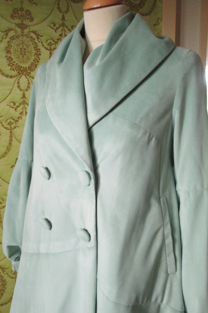 May include: A light blue velvet coat with a large collar and double breasted closure. The coat has a long, flowing silhouette and a single pocket on the right side.