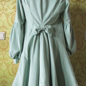May include: A light green velvet coat with a large bow in the back. The coat has long sleeves and a high collar.