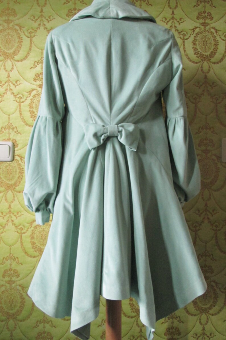 May include: A light green velvet coat with a large bow in the back. The coat has long sleeves and a high collar.