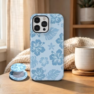 May include: A light blue phone case and matching phone grip featuring a floral pattern of hibiscus flowers and leaves in a darker shade of blue. The phone case is on a wooden surface.