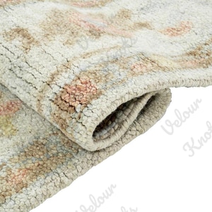 Op de afbeelding: Een dik, handgeknoopt wollen tapijt met een crèmekleurige basis en ingewikkelde bloemmotieven in gedempte tinten beige, zachtroze en lichtblauw. Het tapijt is dubbelgevouwen om de dichte, getextureerde pool en de stevige weving te tonen.