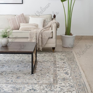 May include: A rectangular area rug featuring a cream-colored base with intricate floral and vine patterns in shades of blue, gray, and beige. The rug has a wide decorative border and a fringed edge, positioned beneath a dark wood coffee table.